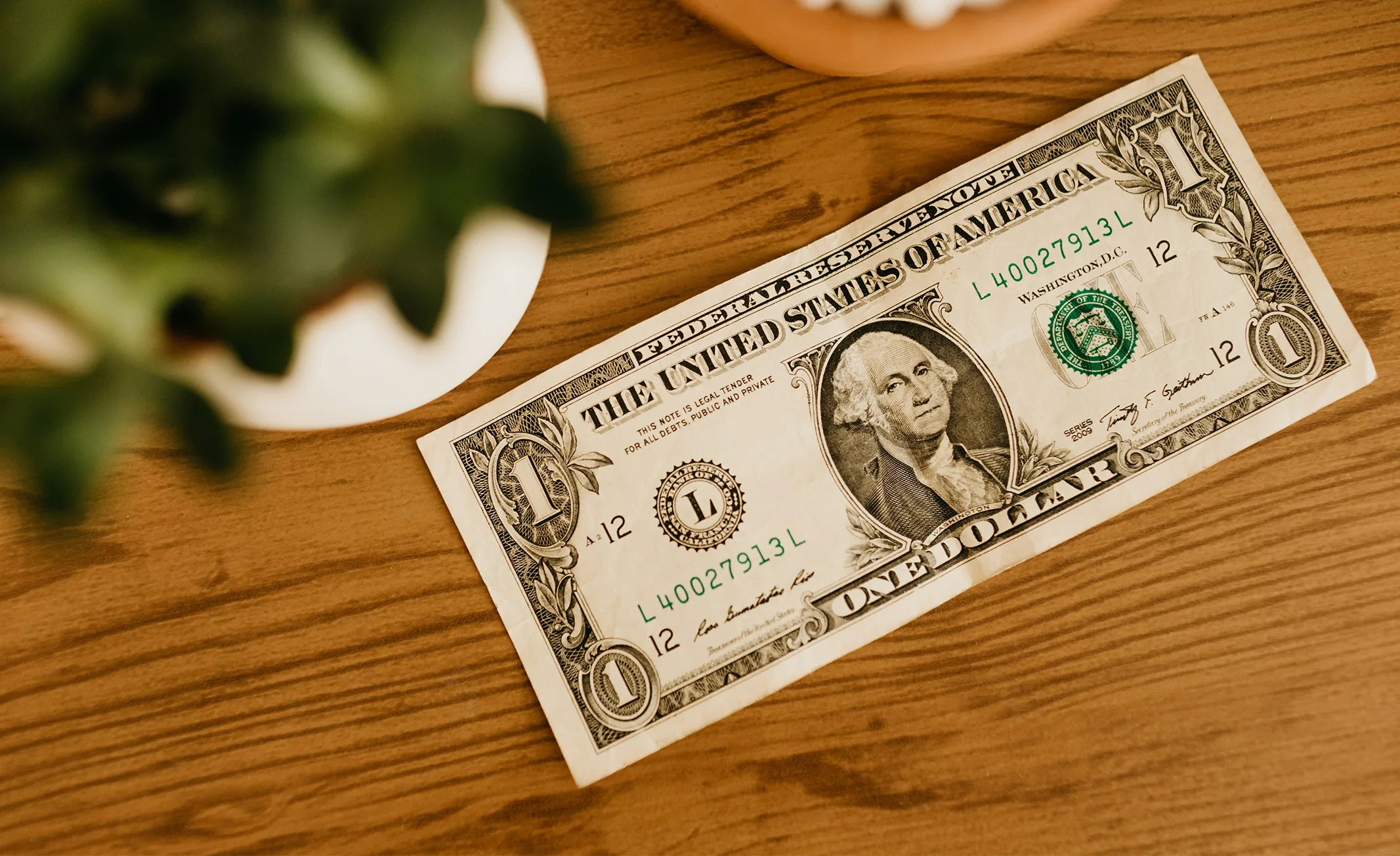A U.S. one dollar bill placed on a wooden table with a blurred plant nearby.